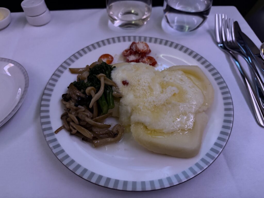 Airline tray table with white table cloth and ceramic dish ware. Served is ravioli with a white sauce, sautéed mushrooms and chilis on the side.