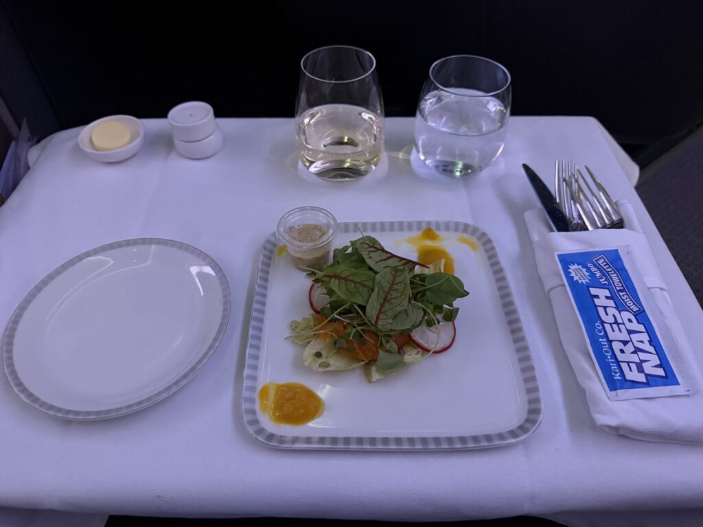 Airline tray table with white table cloth and ceramic dish ware. Served is an arugula salad with an orange sauce on the side.