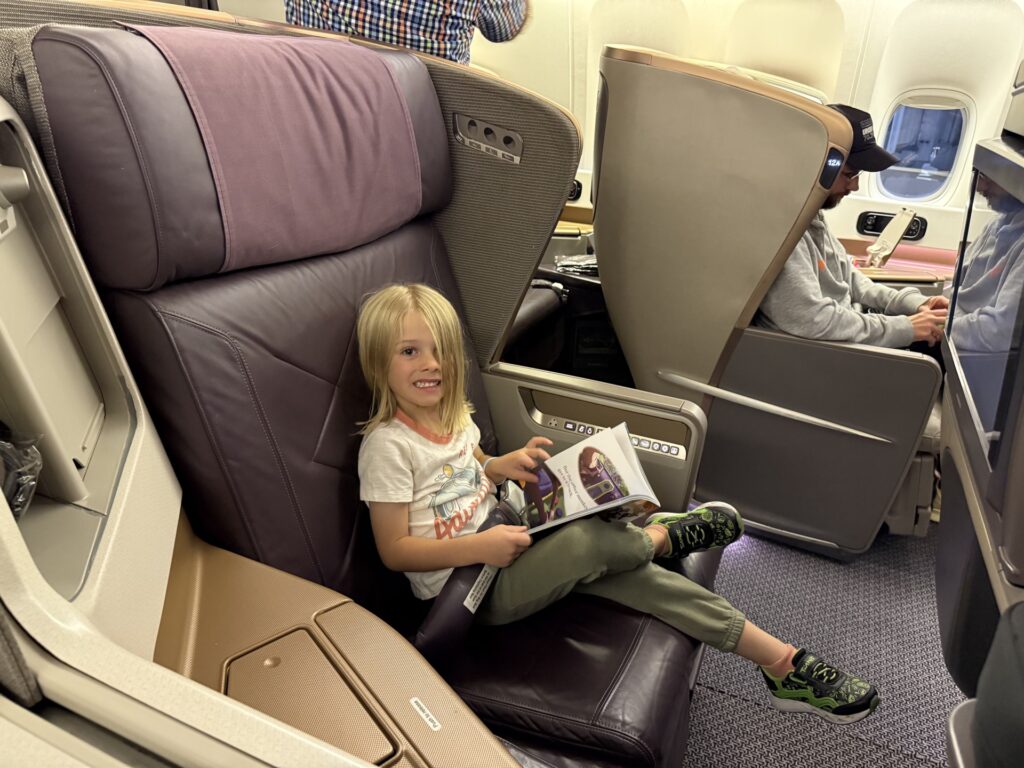 Small child sitting in a large business class seat reading a book and cheesing for a photo.